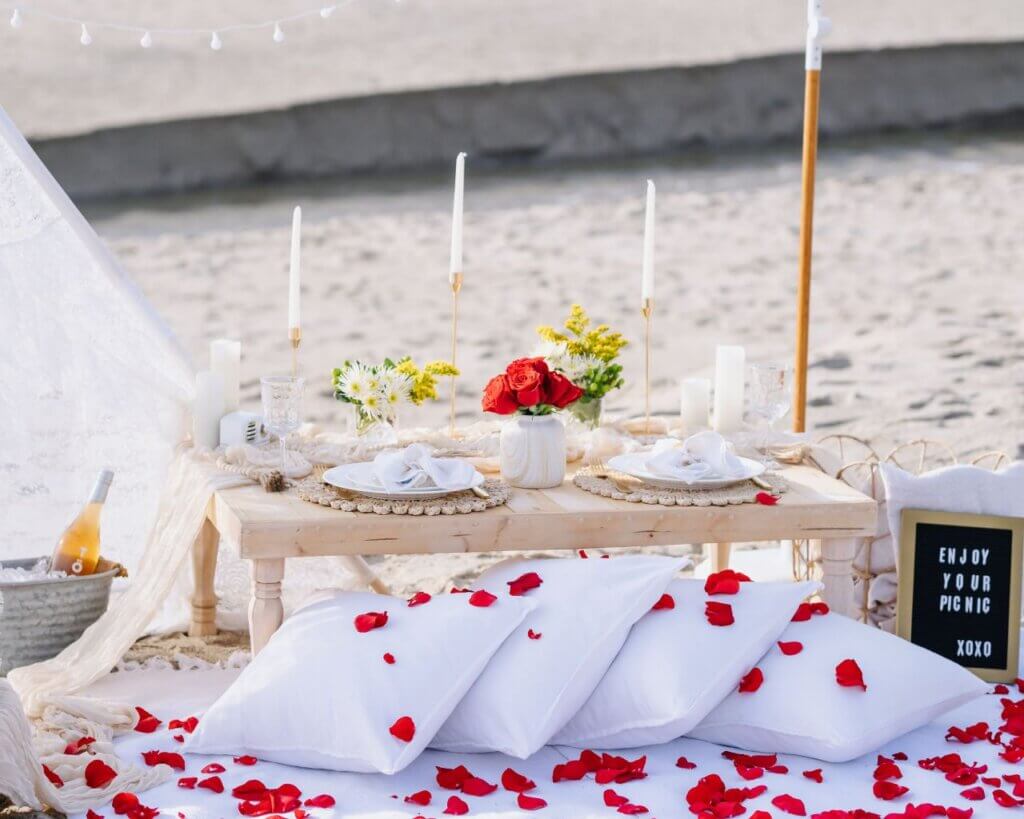 Romantic beach picnic with a white teepee, cushions, candles, flowers, and red rose petals in Malibu