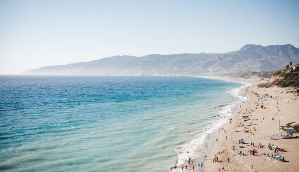 Wide coastal view of clear blue water, sandy shoreline, and mountains in Malibu