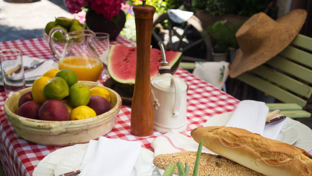Fresh fruit, bread, and flowers styled on a red checkered table cloth for picnic table in a garden setting.