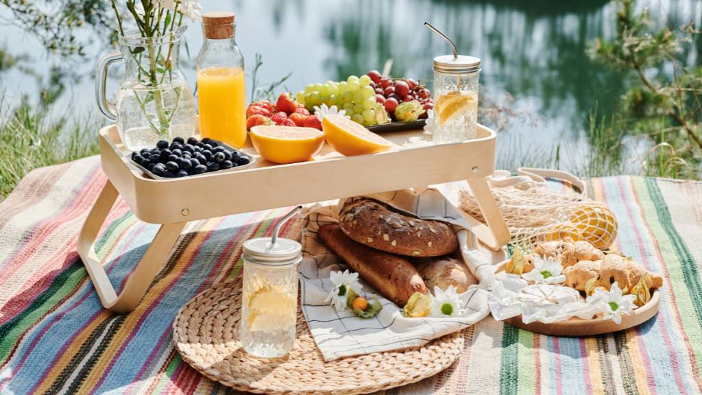 Low picnic table styled with fresh fruit, pastries, and juice on a colorful outdoor blanket near the water.