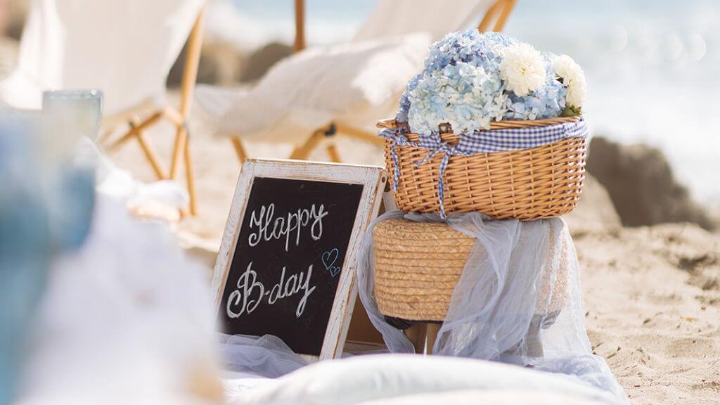 Beach Picnic Birthday Decorations Happy birthday chalkboard beside a wicker basket filled with blue and white flowers at a beach picnic