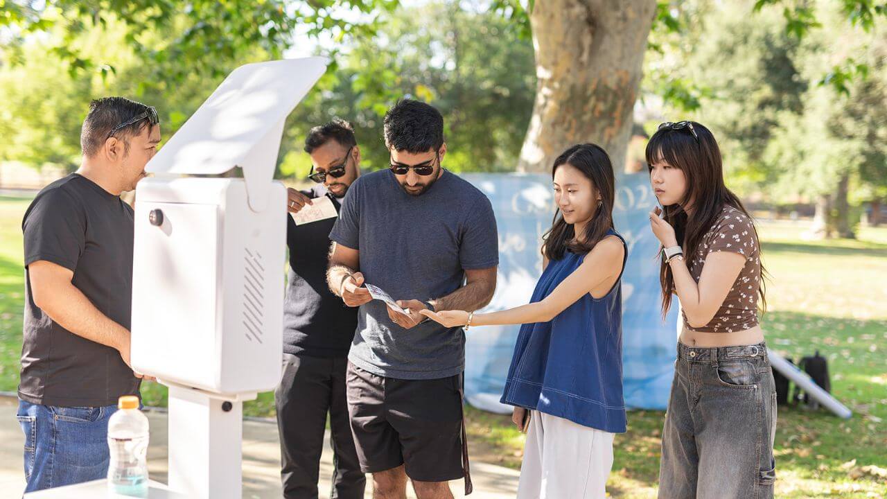 Outdoor Group Photo Booth Activity Group of adults reviewing printed photos beside an outdoor photo booth in a sunny park