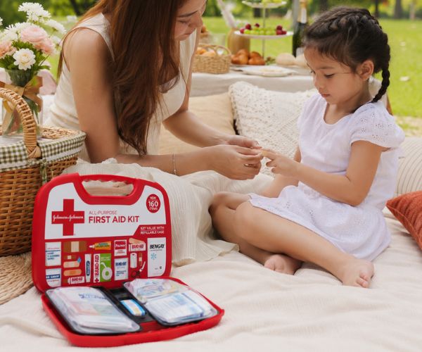 Woman applying a bandage to a child’s finger beside an open Johnson & Johnson compact first aid kit at a park picnic.