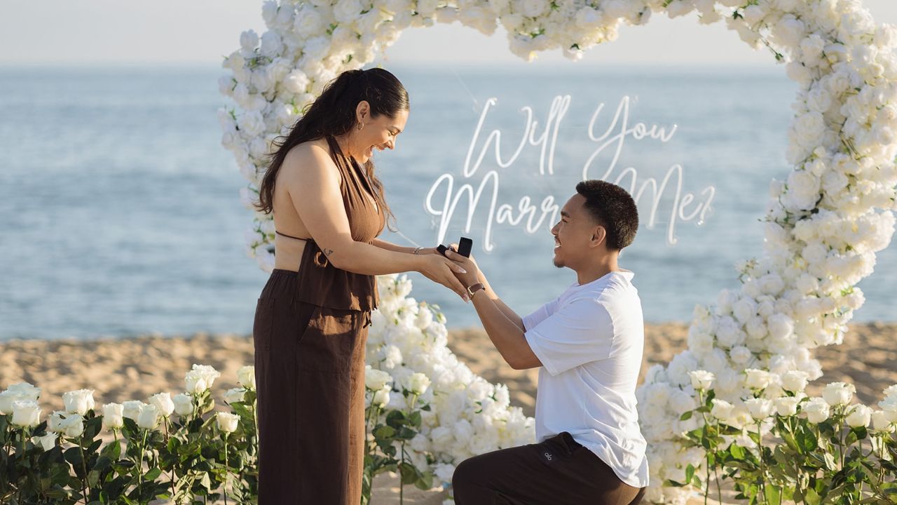 Man kneeling to propose to his partner beneath a white floral arch on the beach with a “Will You Marry Me?” sign