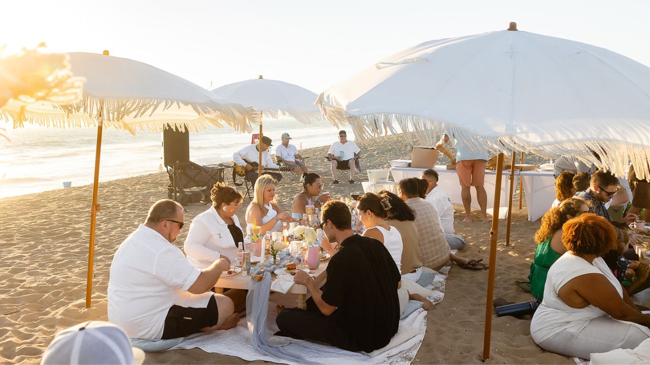 Sunset Group Picnic on the Beach Large group enjoying a sunset beach picnic at low tables beneath white fringed umbrellas