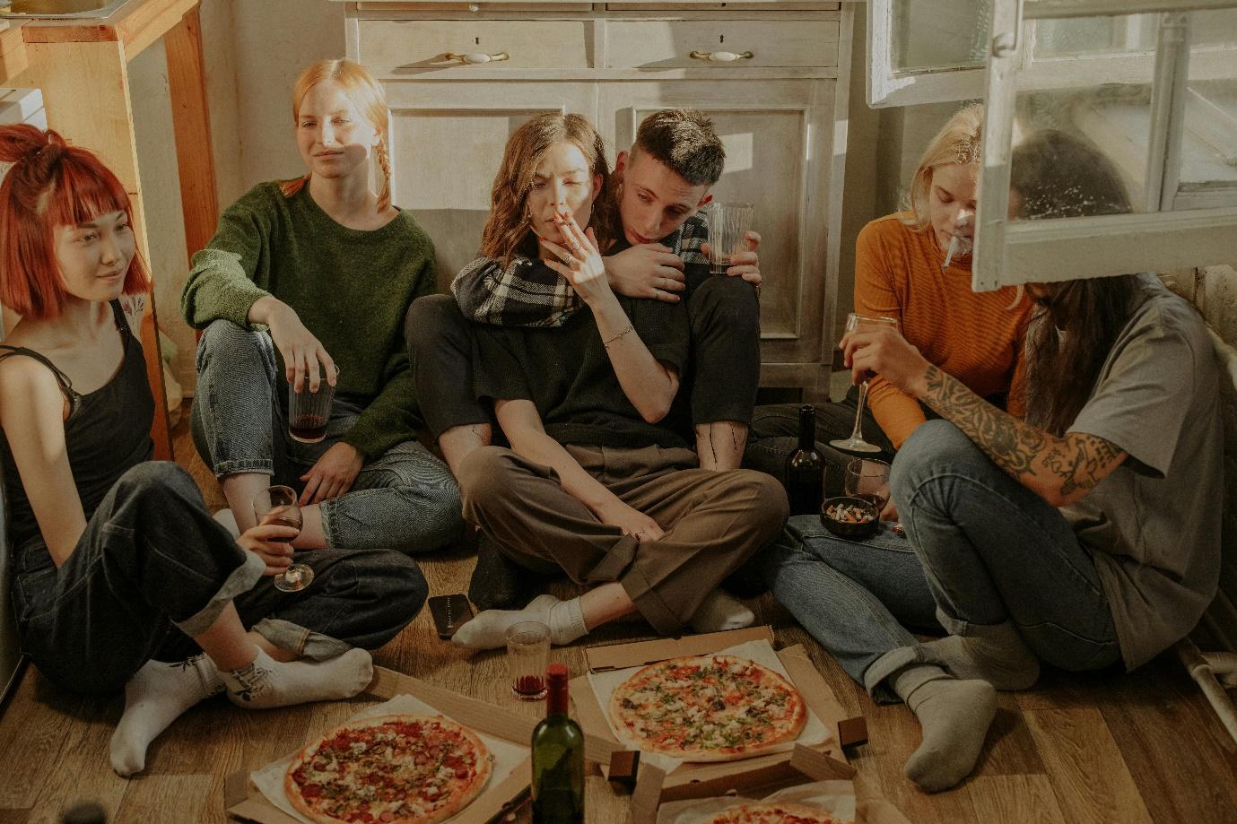 Group of friends sitting on the floor sharing pizza and drinks at a casual gathering.