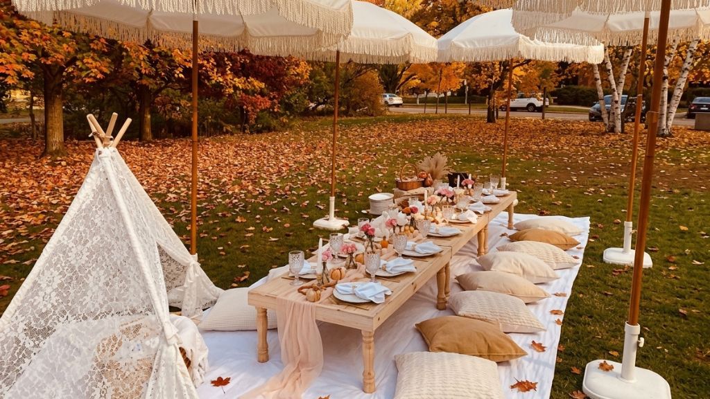 Fall picnic setup with low tables, white cushions, teepees, and umbrellas beneath autumn trees