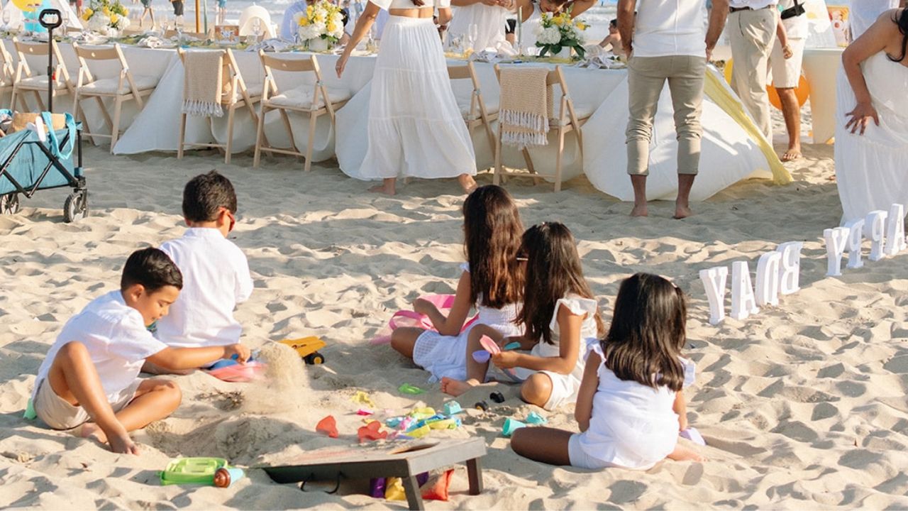 Children playing in the sand during a beach birthday party with decorated tables and guests in the background